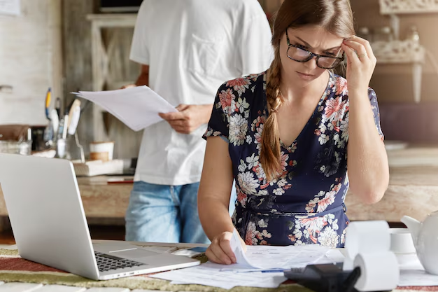 Woman in glasses studying paperwork at a laptop while a man reads documents behind her